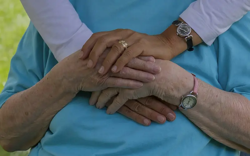 A support worker in a blue top gently holds an older person's hands