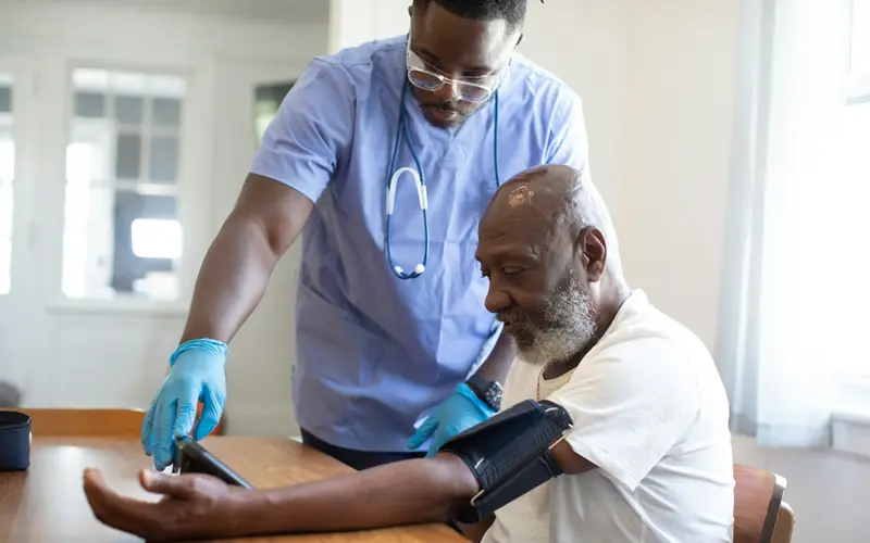 A support worker in gloves checks the blood pressure of a seated man at a table at home